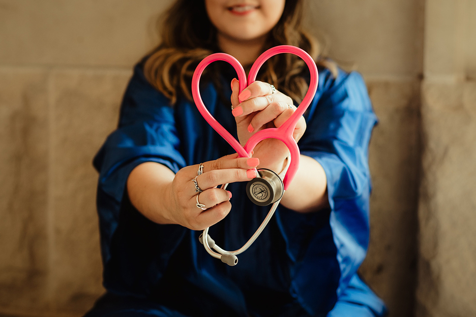 Nurse in blue scrubs holding up a pink stethoscope in the shape of a heart.